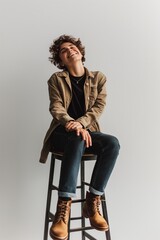 Happy young person with curly hair seated on a stool, laughing and looking carefree, complimented by casual fashion and neutral backdrop. - obrazy, fototapety, plakaty