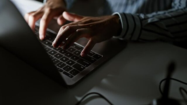 Close-up of a man's hands typing on a laptop keyboard in a dark office setting, depicting work and technology