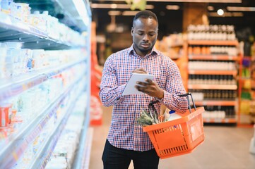 african american man standing in supermarket choosing products while reading notes at notebook