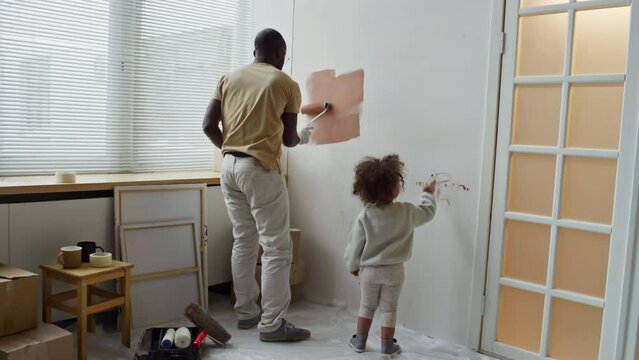 Rear full footage of father painting drywall in brown with paint roller and little curly-haired baby helping him during room renovation