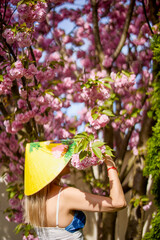 portrait of a blonde woman with a Japanese hat next to a blooming cherry tree