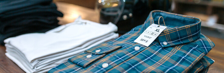 Close up of label with price and size over a blue plaid shirt on industrial style store. Banner of...