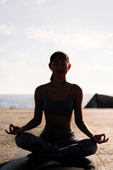 Fototapeta premium backlight of young woman doing meditation by the sea sitting with legs crossed, concept of mental relaxation and healthy lifestyle
