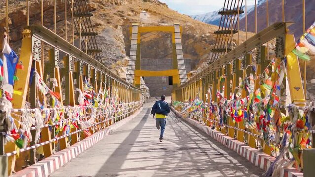 Slow motion shot of an Indian male tourist running on the Chicham bridge with prayer flags waving on its side at Chicham in Spiti Valley, Himachal Pradesh, India. Tourist running on a bridge in Spiti.
