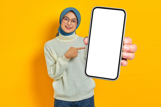 Portrait of cheerful young Asian Muslim woman in white sweater and hijab pointing to blank mobile phone screen isolated on yellow background. Muslim lifestyle concept