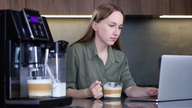Woman drinking coffee and working on laptop near coffee machine at home.