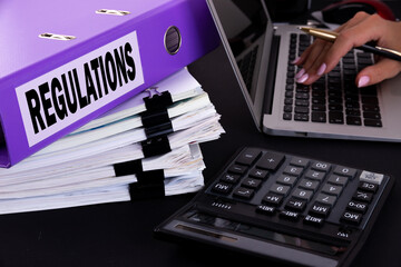 Text, word Regulations is written on a folder lying on documents on an office desk with a laptop...