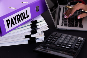 Text, word Payroll is written on a folder lying on documents on an office desk with a laptop and a...