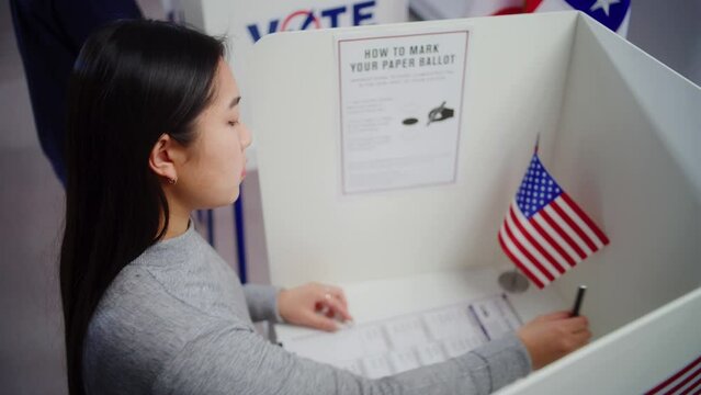 Asian woman comes to voting booth at polling station, makes choice and fills out paper bulletin. US citizen during National Election Day in the United States of America. Civic duty and patriotism.