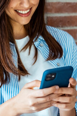 Young modern Caucasian girl smiling looks at cell phone, standing leaning against brick wall. Relaxed female looks at the phone. Technology concept.