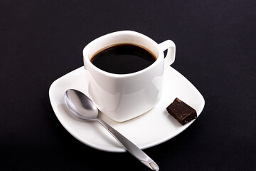 White cup with coffee on a saucer with a spoon and a bar of chocolate on a black table.