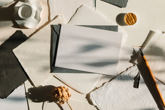 Overhead view of a blank piece of homemade deckle edge card, calligraphy pen, envelope and flowers on a table in sunlight