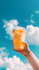 Hand holding clear plastic cup with cold refreshing orange beverage and a blue straw against cloudy sky.. Summer background.