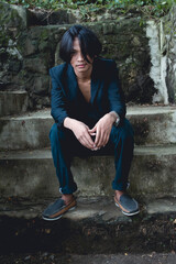 Young Asian man in goth-inspired attire sitting solemnly in a forest setting