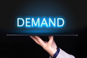 Man hand holds a tablet over which a neon text is written, the word DEMAND. Business concept.