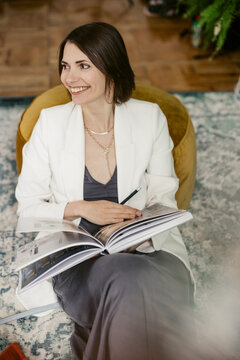 Brunette girl  in white 
jacket reading book and smil in room with green wall