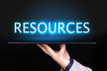 Man hand holds a tablet over which a neon text is written, the word RESOURCES. Business concept.