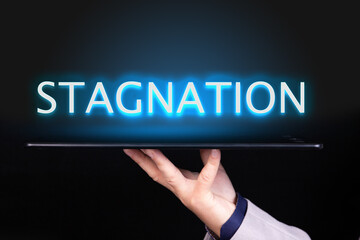Man hand holds a tablet over which a neon text is written, the word STAGNATION. Business concept.