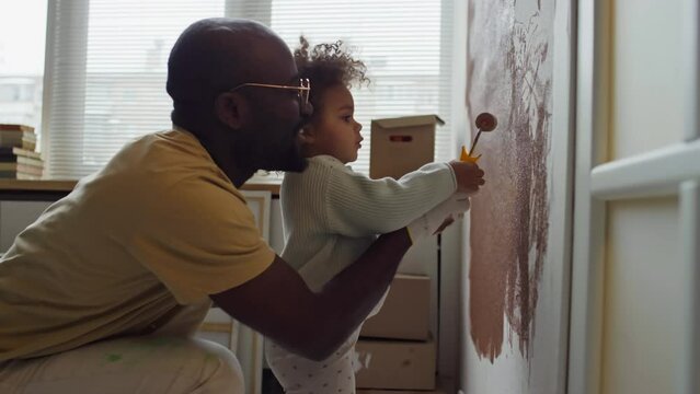 Side footage of caring African American father helping his Biracial child learn to paint walls using paint roller during home redecoration
