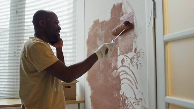 Side footage of Black man making phone call while painting wall brown using paint roller in new apartment
