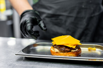 Chef in black attire garnishing a freshly made burger with cheddar cheese on a metal tray, showcasing the precision in food preparation