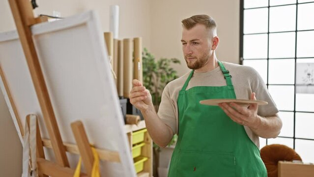 A focused man artist in a green apron paints on a canvas in a bright studio
