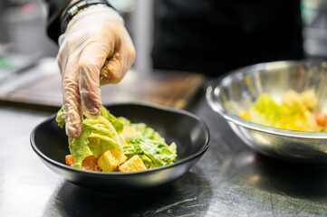 Chef’s gloved hand meticulously placing fresh salad into a black bowl, emphasizing hygiene and precision in culinary arts