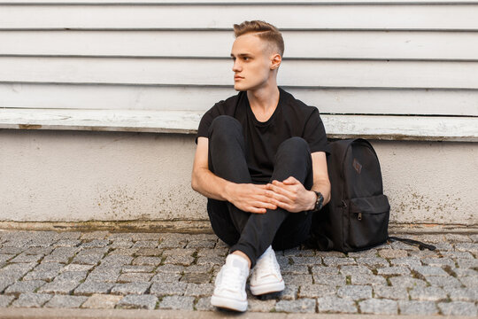 Stylish student handsome guy with a hairstyle in fashionable black clothes with a T-shirt, jeans and white sneakers with a backpack sits near a wooden wall
