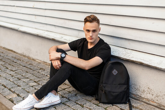Handsome fashion guy model with a hairstyle in black clothes with white sneakers and a backpack sits on the street near a wooden wall