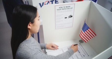 Asian woman comes to voting booth at polling station, makes choice and fills out paper bulletin. US...