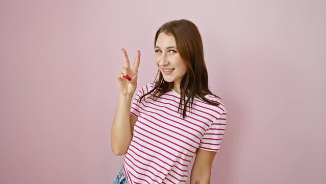 Joyful brunette girl in striped t-shirt flashing victory sign, beaming with win-win confidence, over pink isolated backdrop