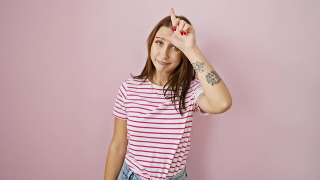 Cheeky young brunette girl in striped t-shirt, poking fun at people by flashing the loser sign with her fingers on her forehead, insolently mocking over isolated pink background.