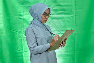 beautiful asian woman wearing hijab, glasses and blouse seriously writes something in the folder...