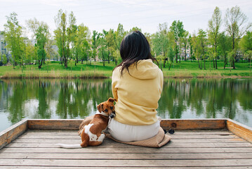 Woman sits on a pier with a Jack Russell dog near a pond in the park. Dog is sitting. He turns around and looks at the camera.	