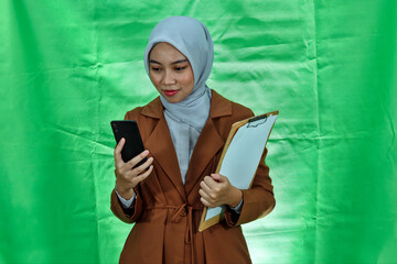 beautiful asian wearing hijab and blazer woman holding clipboard and mobile phone on green...