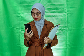 beautiful asian wearing hijab, glasses and blazer woman holding clipboard and mobile phone on green...