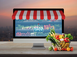 Full fresh food in shopping basket and supermarket in computer monitor screen with online shopping store graphic and open sign on wooden table over blur of cityscape on warm light sundown