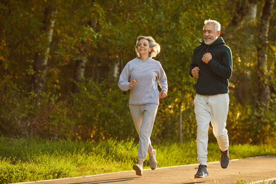 Retirees wife and husband rejoice in active lifestyle. Lovely joyful retirees couple doing sports and jogging together in city park. Senior man and woman in sportswear running on asphalt path.