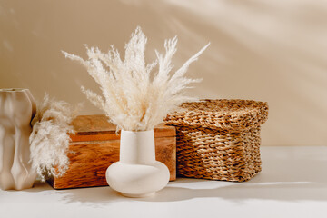 Bohemian interior still life composition. Ceramic vase with pampas grass, wooden box and wicker storage container with sunlight shadows. Scandinavian home interior decorations for poster.