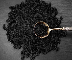 Black Caviar in golden spoon on caviar backdrop. High quality natural sturgeon black caviar close-up. Slate stone background. Delicatessen. Texture of expensive luxury caviar, top view.