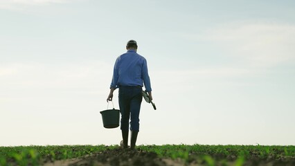 Farmer with shovel bucket walks through agricultural field. Tired exhausted man male businessman...