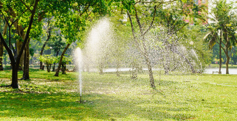 Water sprinkler system in action, watering green grass in a park with sunlight filtering through...