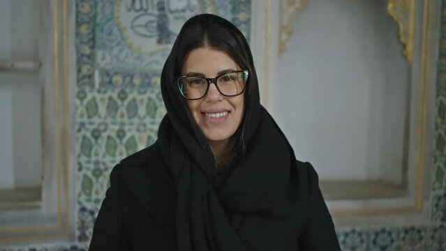 A smiling young woman with glasses, wearing a black shawl, stands in the ornate topkapi palace in istanbul.