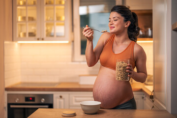 A satisfied pregnant woman holding jar of cereal and eating oatmeal for breakfast in the kitchen