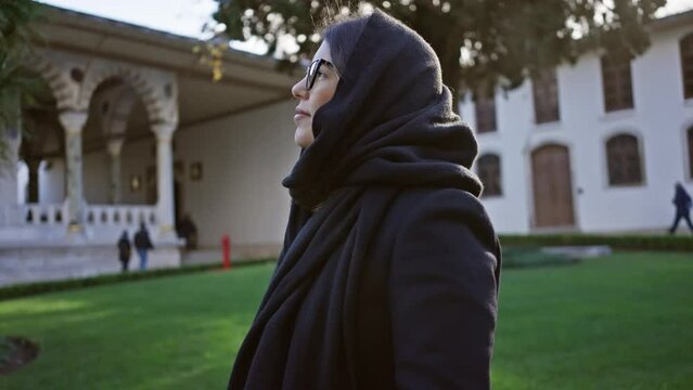Smiling woman with glasses and headscarf at historic topkapi palace in istanbul, leisurely experiencing turkish culture and architecture.