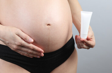Pregnant woman in underwear holding cream tube on gray background