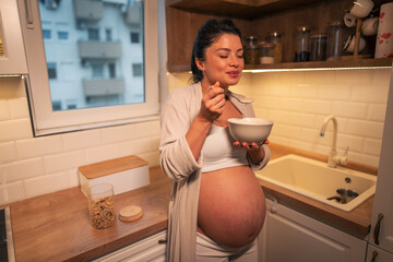 A contended pregnant woman in pajamas eating a bowl of cereal for breakfast in the kitchen