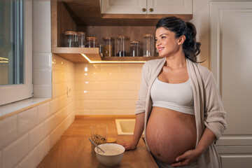 A beautiful adult pregnant woman having breakfast in the kitchen and looking outside the window