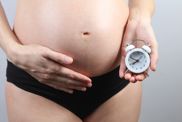 Pregnant woman in underwear holding alarm clock on gray background