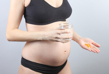 Pregnant woman in underwear holding fish oil capsules and water glass on gray background